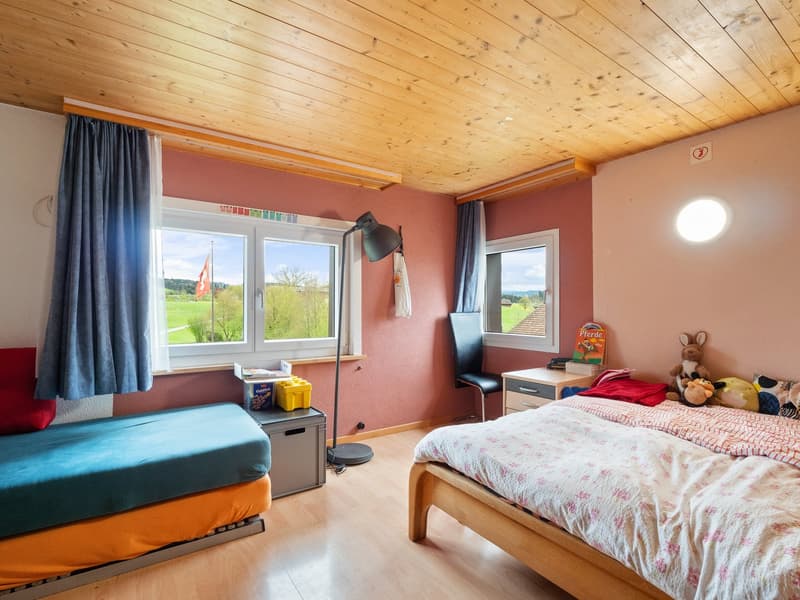 Spacious bedroom with wooden ceiling, pink walls, large windows overlooking a scenic outdoor view, a bed, a sofa, and various personal items and decor.