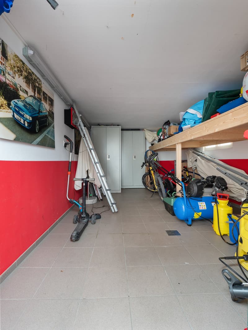 The image shows a cluttered garage space with various items stored on shelves and the floor. There is a bicycle, a ladder, and other tools and equipment visible. The walls are painted in a red and white color scheme, and the floor is tiled.