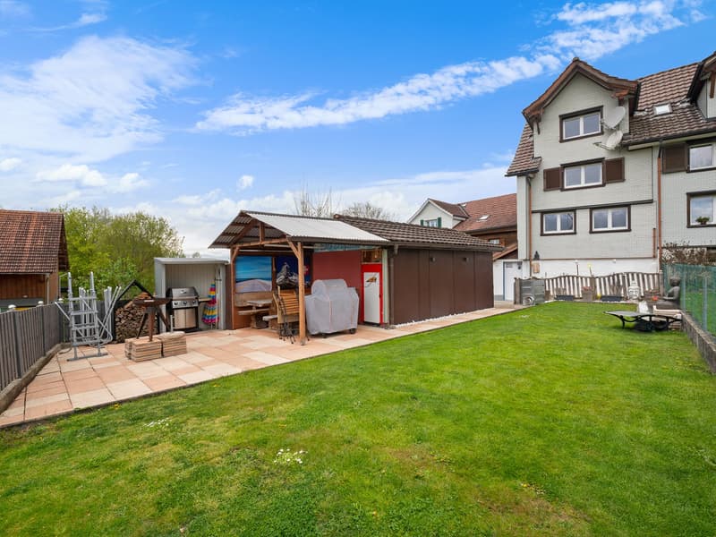 Two-story residential building with a tiled roof, surrounded by a grassy yard and a paved patio area. The yard features a wooden shed or storage structure, a barbecue grill, and other outdoor furniture. There is also a garage or carport visible on the pro
