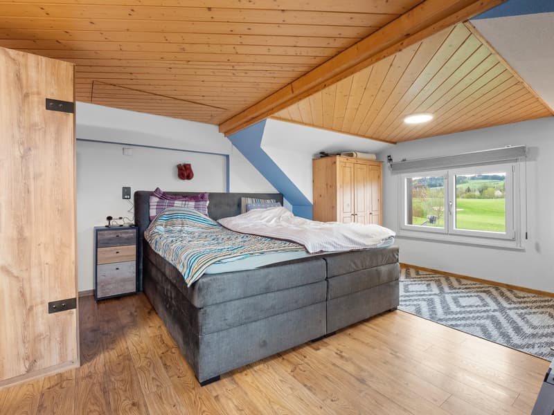 Spacious bedroom with wooden ceiling and walls, large window overlooking a scenic outdoor view, gray upholstered bed, wooden wardrobe, and area rug on hardwood floor