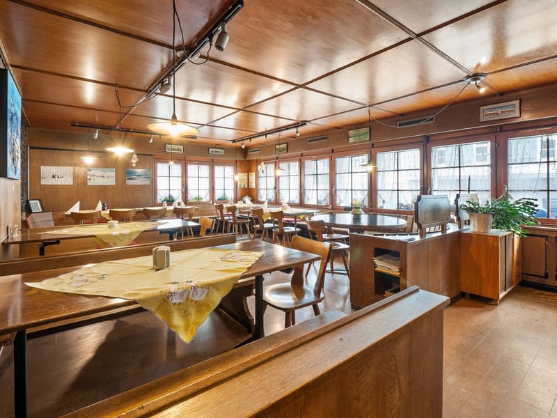 Spacious dining area with wooden walls, ceiling, and furniture. Large windows provide natural lighting. Tables are set with tablecloths and there is a counter with a dishwasher visible.