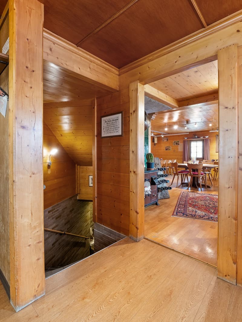 Wooden cabin-style interior with exposed wood beams and paneling, hardwood floors, and a framed wall decor. The room opens up to a larger living and dining area with a rug and furniture visible through the doorway.
