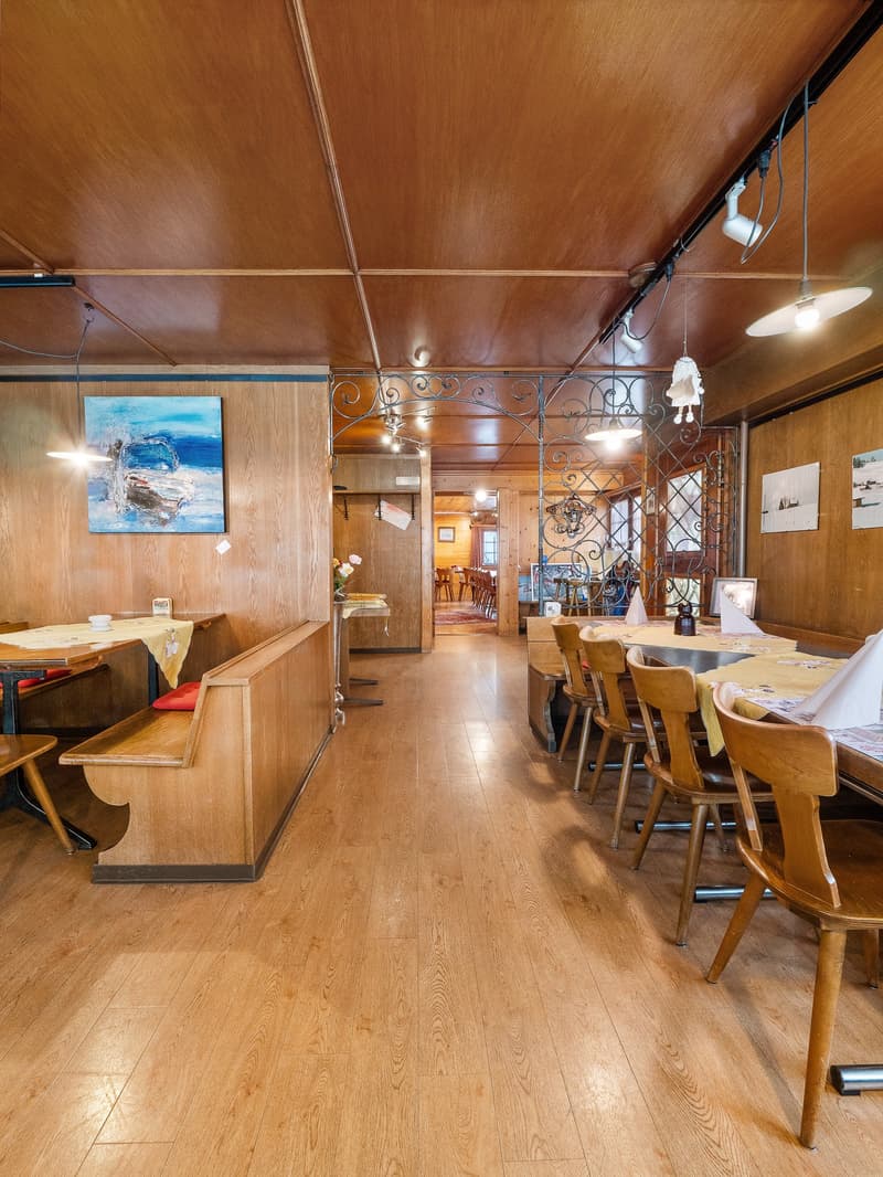 Cozy dining area with wooden walls and ceiling, ornate metal light fixtures, artwork on the walls, and a fireplace visible in the background.