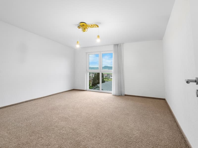 Empty room with carpeted floor, white walls, large glass door leading to a view of a town, two yellow lamps hanging from the ceiling.