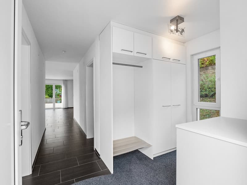 A white hallway with tile floor and carpeted floor on one side, white cabinets, a closet with hanging rod, ceiling lamp, and windows overlooking a garden