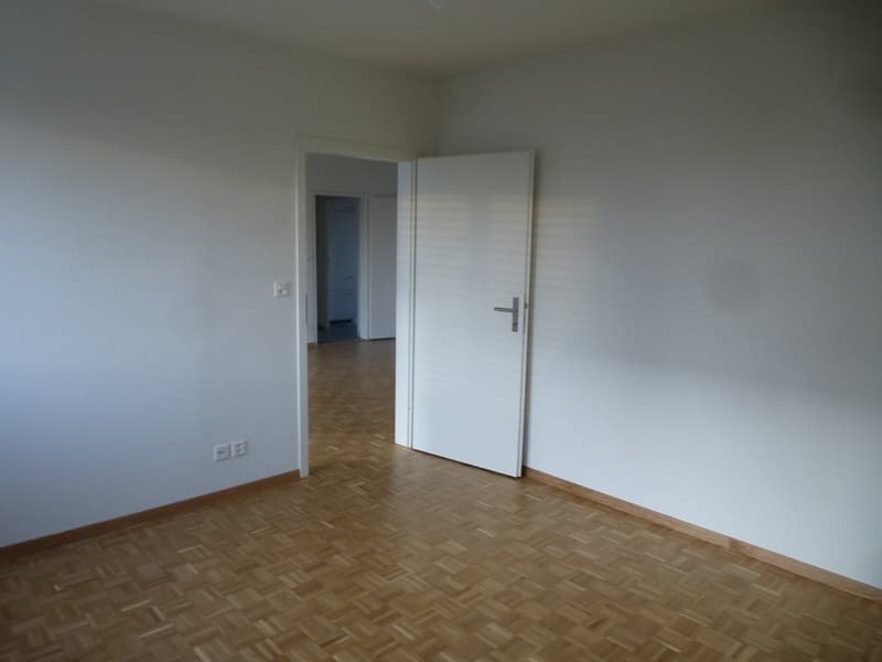 An empty room with wooden floors, white walls, a white door, and an open door leading to another room with a white cabinet