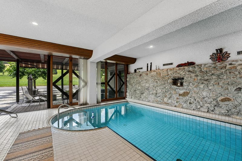 Indoor swimming pool with wooden deck, adjacent stone wall, and glass doors leading to an outdoor patio.