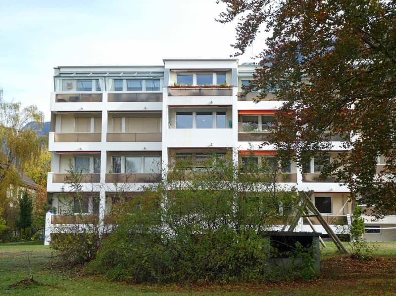 3-story apartment building with white exterior, large windows, and balconies. The building is surrounded by trees and greenery.