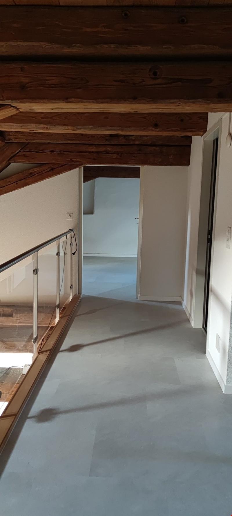 The image shows the interior of a room with a wooden ceiling and exposed beams. The floor appears to be a light-colored tile or laminate. There is a doorway visible in the background, suggesting this may be a hallway or entryway leading to another room.