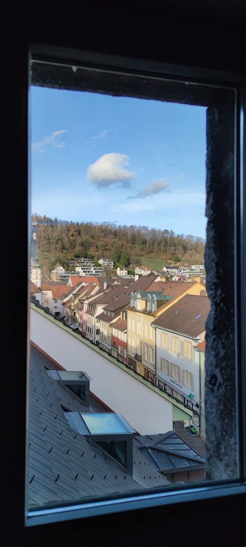 View of a quaint European town with colorful, multi-story buildings, a forested hillside in the background, and a cloudy blue sky above.
