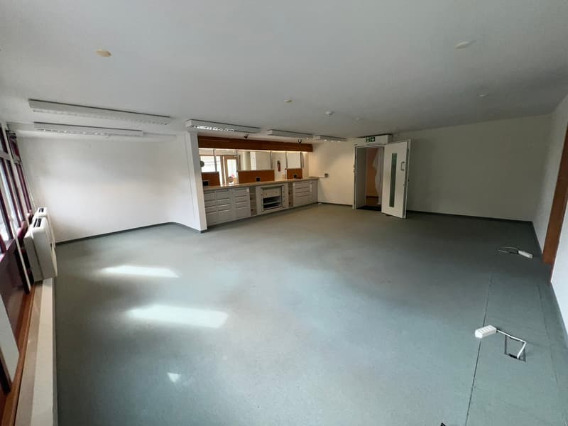 Large open-plan room with white walls, concrete flooring, and built-in kitchen cabinets and appliances. The room appears to be a combination of a living area and a kitchen space.