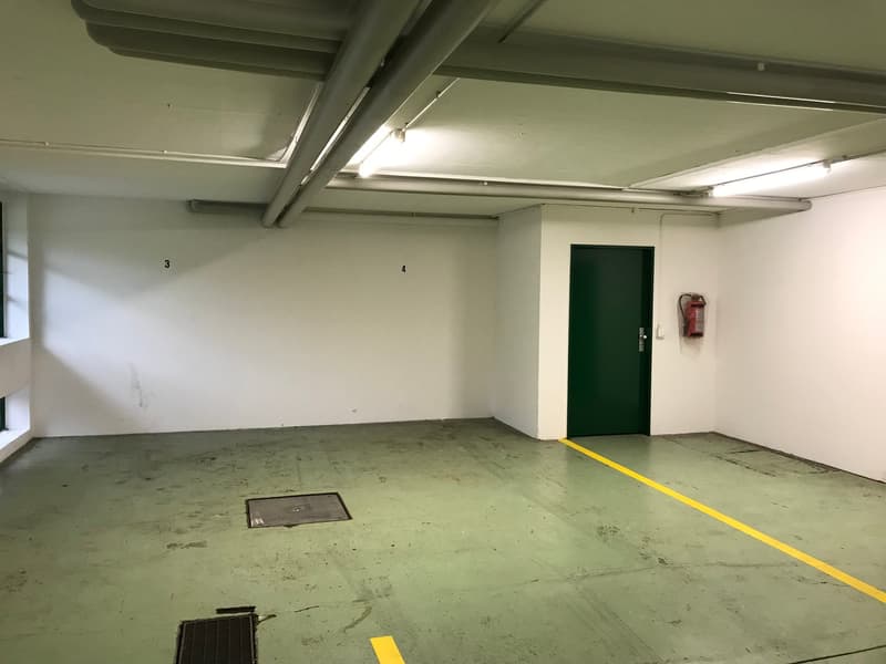 Spacious garage with concrete floor, exposed ceiling beams, and a green door leading to another room or area.