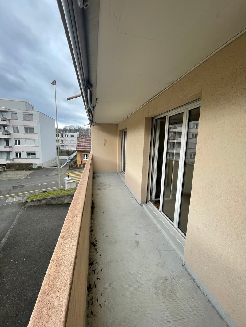 Balcony of a building with glass sliding door and railings, paved floor, beige walls, street, building in background, street light