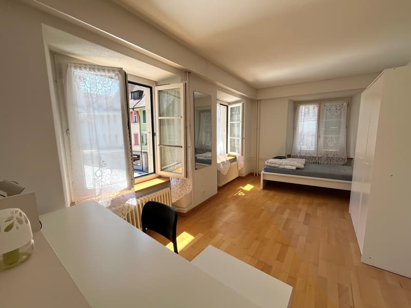 wooden floor, white walls, open windows, bed, desk, chair, white curtains, radiator, small decorative object on desk
