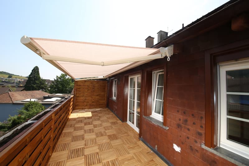 The image shows a wooden balcony or terrace attached to the side of a building. The balcony has a retractable awning or canopy that is extended, providing shade. The balcony floor is made of wooden tiles or panels. The building itself appears to be a trad