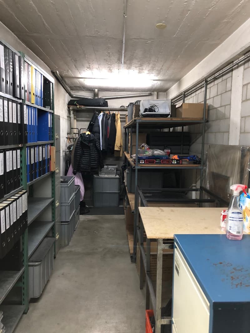 Store room with shelves, storage boxes, shelves with files, clothing hanging, tools on table, boxes, plastic cover, storage box, spray bottle, paper on wall