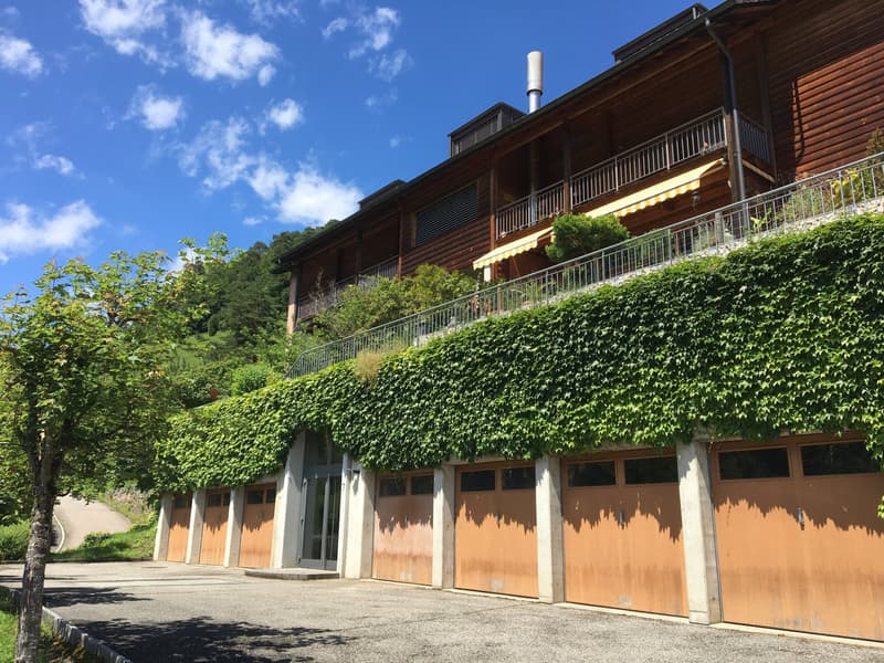 a wooden building with multiple garage doors and a balcony on top, surrounded by greenery
