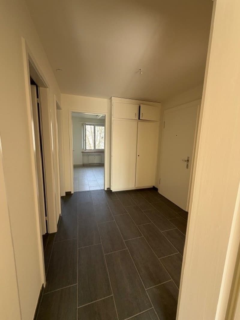 Empty hallway with white walls, wooden floor, white cabinets, and a window.