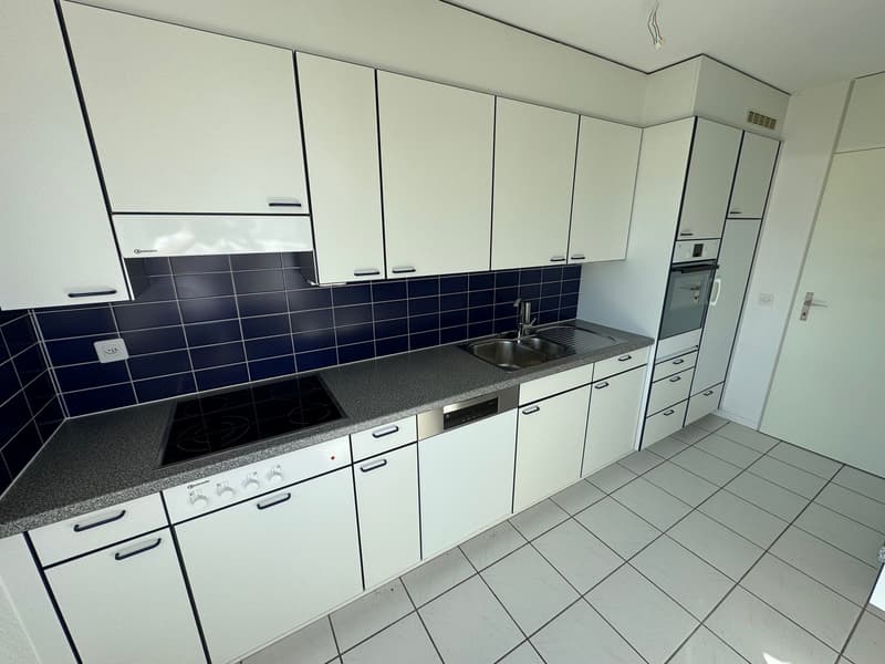 modern white kitchen with blue backsplash tiles, stainless steel sink, white tiled floor, black induction hob, integrated dishwasher, hanging cabinets, and built-in oven.
