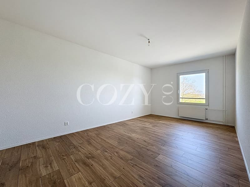 Spacious bedroom with white walls, hardwood flooring, and a large window providing natural light.