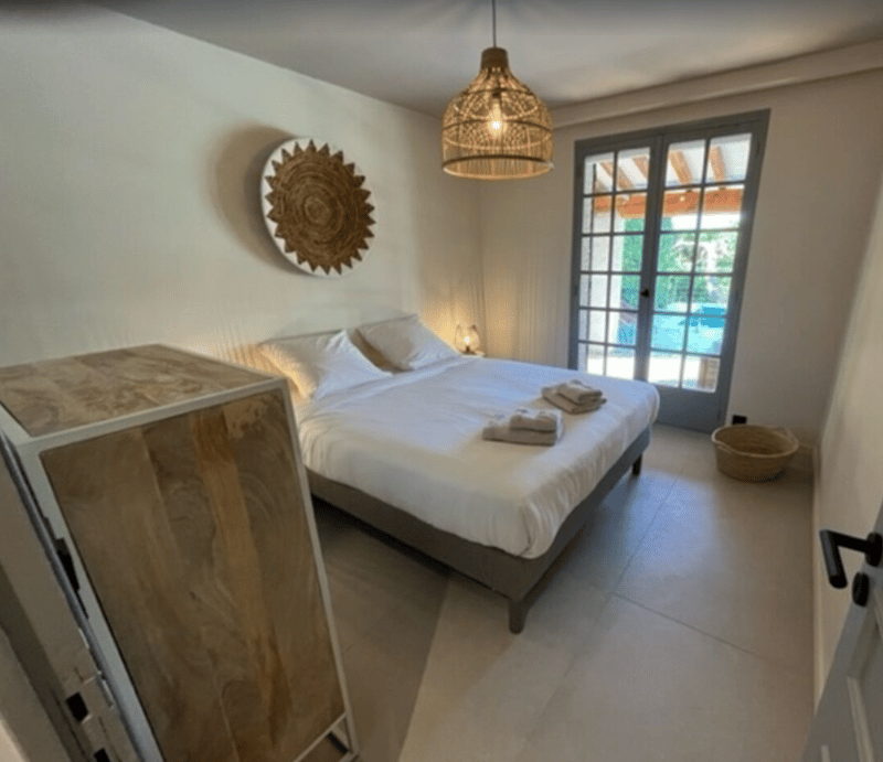Bedroom with white walls, bed with white sheets, two white pillows, decorative basket, sliding door with pool view