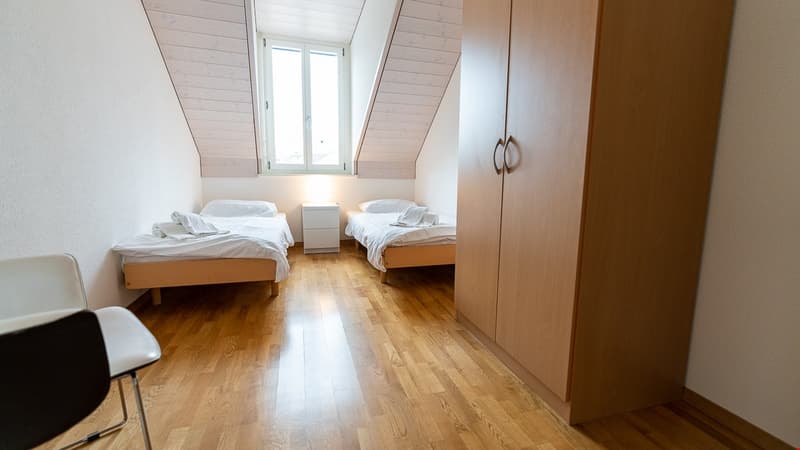 2 beds, wooden floor, white walls, slanted ceiling, window, bedside table, chair