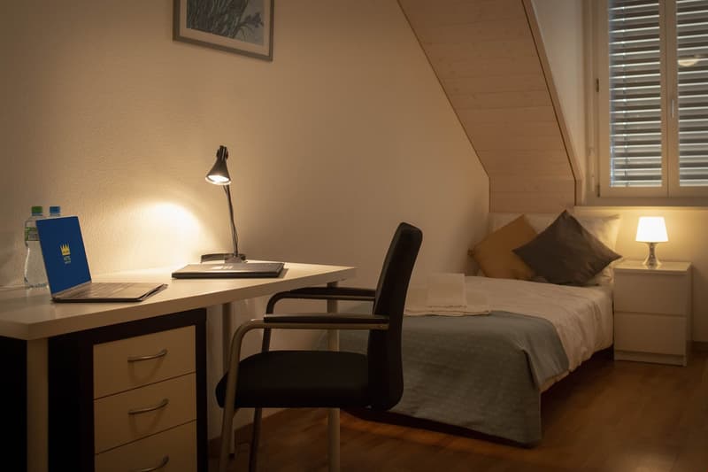 small bedroom with bed, desk, lamp, chair, bottle, frame, window, wooden floor