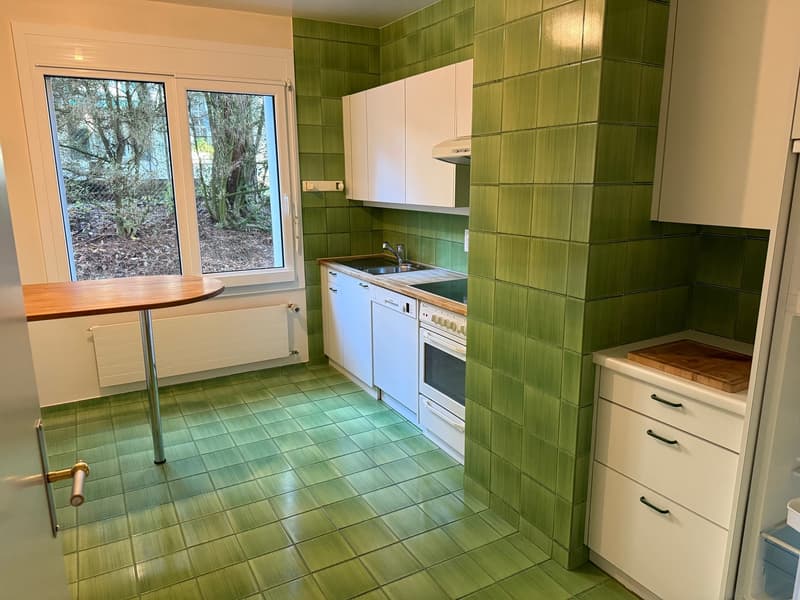 Green tiles on the walls and floor, white cabinets, sink with faucet, dishwasher, oven, a small round table, and a window with trees outside.