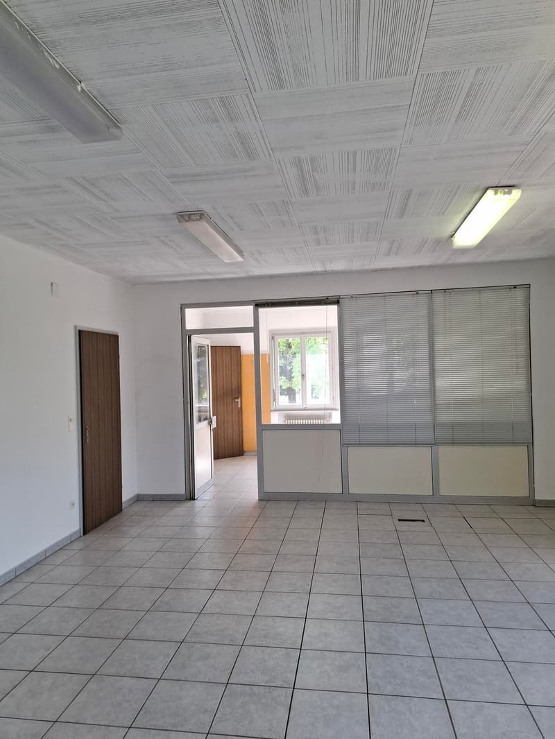Spacious room, gray tile floor, white walls, closed doors, ceiling lights, open door to adjacent room with glass door and window, and a white partition with blinds.
