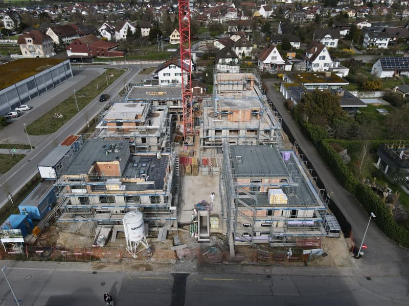 Aerial view of a construction site with multiple buildings under construction, a large red crane, and various construction materials and equipment scattered around.