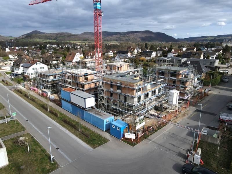 Aerial view of a construction site with multiple buildings under construction, a crane, and surrounding houses and landscape.