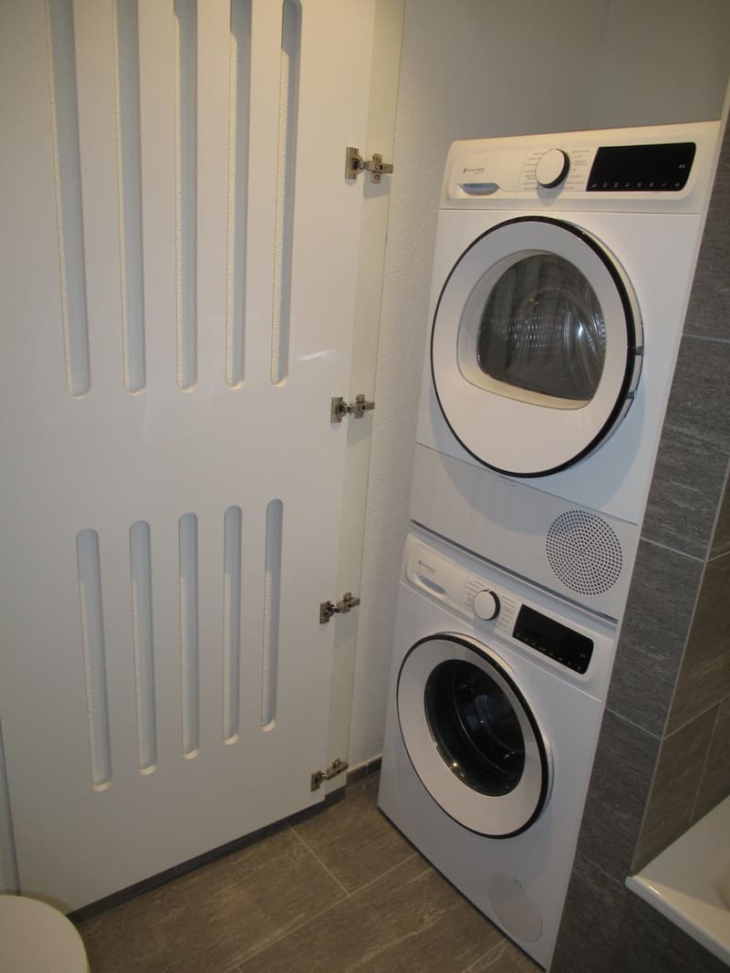 A white door with vertical stripes, a white washing machine and dryer stacked on top of each other, a white toilet, and a white sink.