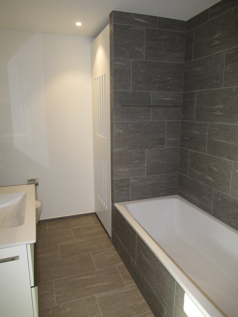 Bathroom with bathtub, sink, and toilet, gray tiled walls, white cabinets, wooden floor