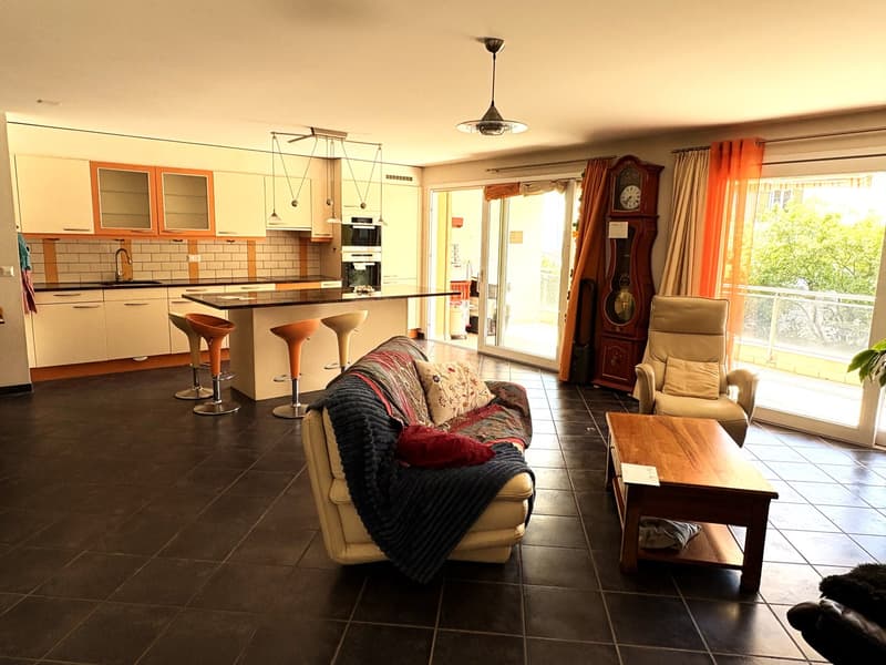 Open plan living room and kitchen, large windows, balcony, wooden table, couch, armchair, grandfather clock, kitchen island, stools, cabinets, oven, sink