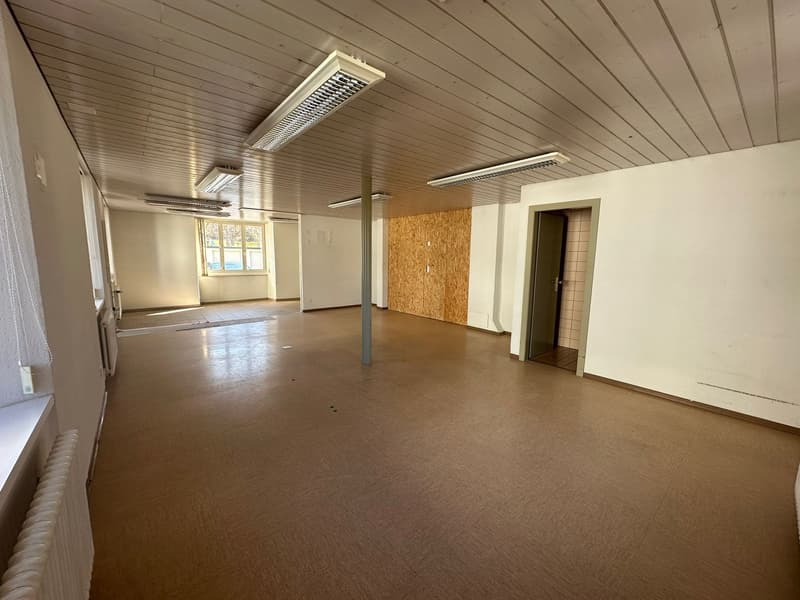 Empty room with wooden floor, white walls, ceiling with wooden beams and lights, open door, window with white curtains, radiator