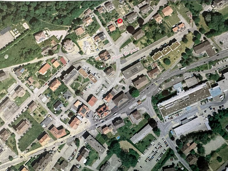 Aerial view of a town with multiple buildings, roads, and parking areas. The area is surrounded by trees and grass.
