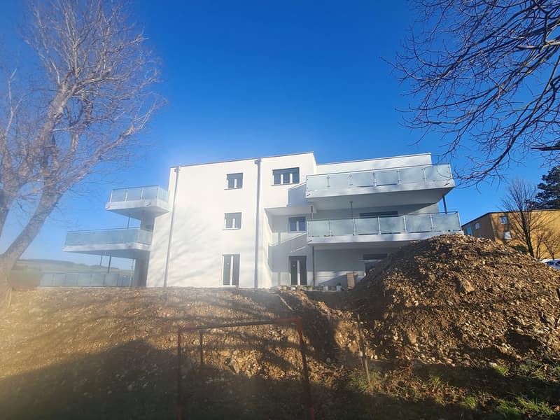 Modern white building, 3 stories, large windows, multiple balconies, trees, pile of dirt in front, clear blue sky, distant house
