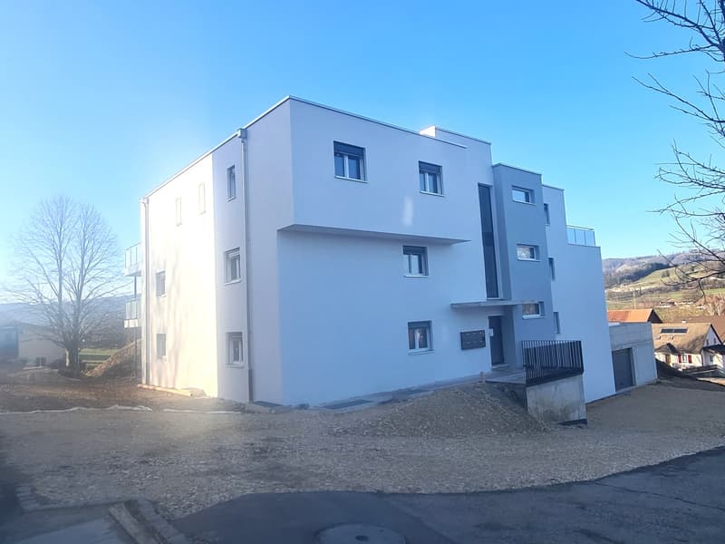 Three story white residential building, several windows, surrounded by construction material and a barren landscape, mountains in the distance