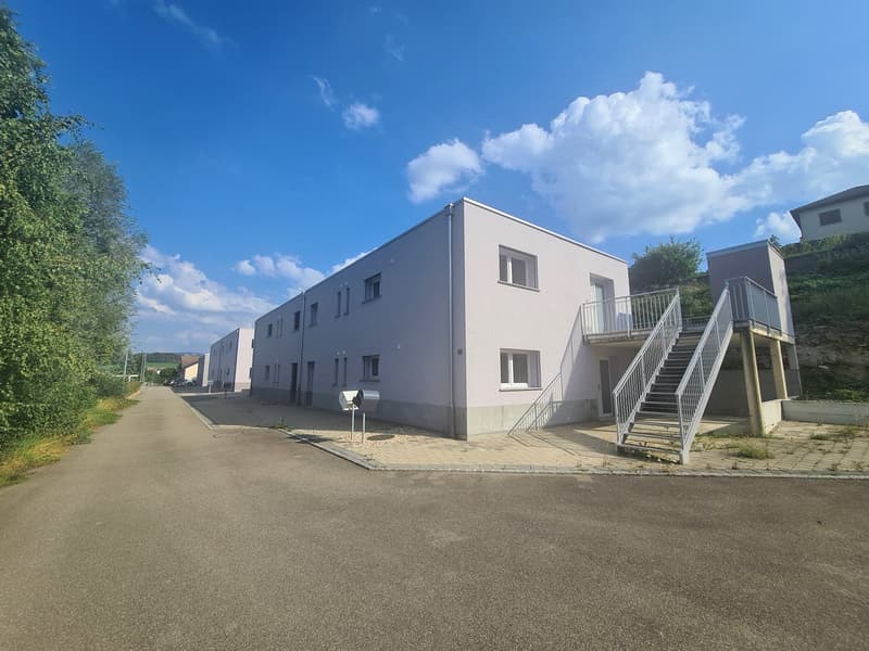 Modern apartment building, light gray facade, white windows, concrete pathway, small staircase, metal railings, large window on first floor