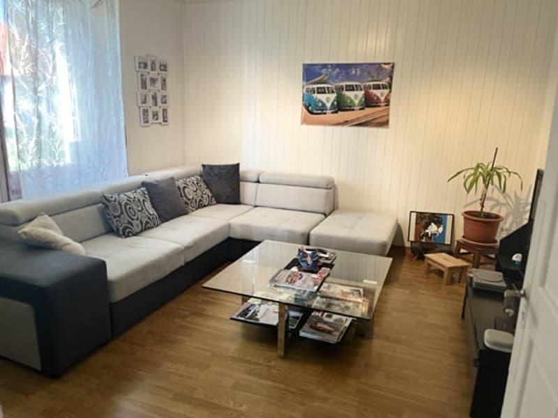 Spacious living room with a large L-shaped sectional sofa, a glass coffee table with magazines, and a wall-mounted TV. The room has wooden flooring and a curtained window, suggesting the possibility of a balcony or terrace.