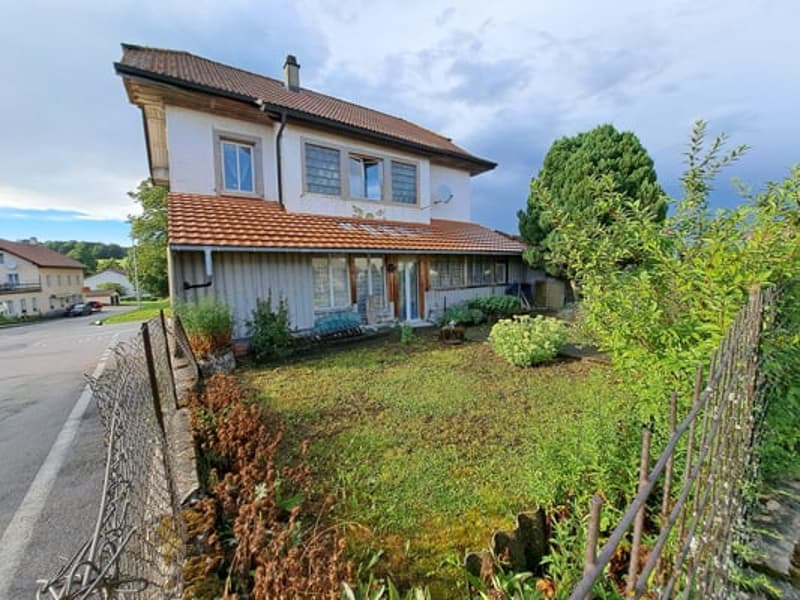 2-story wooden house with a tiled roof, surrounded by a grassy yard and trees. There is a balcony or terrace visible on the upper floor, and a parking space or driveway in front of the house.