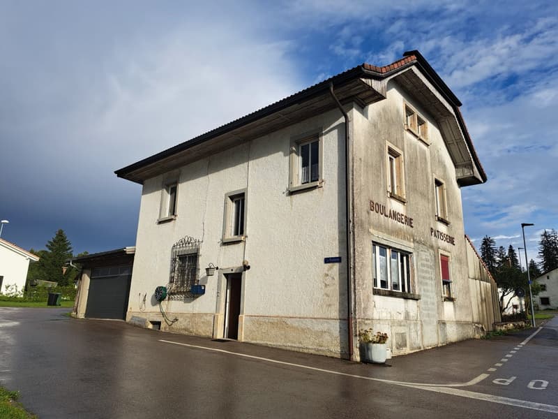 A two-story building with a slanted roof, white exterior walls, and several windows. The building has a sign that says 'Boulangerie Patisserie', indicating it is a bakery or pastry shop. There is a garage door on the side of the building.