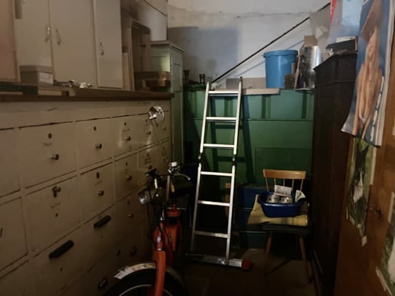 The image shows a cluttered storage room or basement area. There are various items and boxes stored on shelves and the floor, including a ladder, a fire extinguisher, and some plastic containers. The room appears to be dimly lit and has a utilitarian, unf