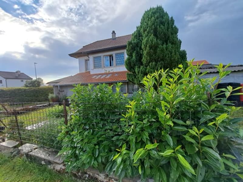 Two-story house with a red tile roof, surrounded by lush greenery and vegetation. The house has large windows and appears to have a balcony or terrace on the upper level.