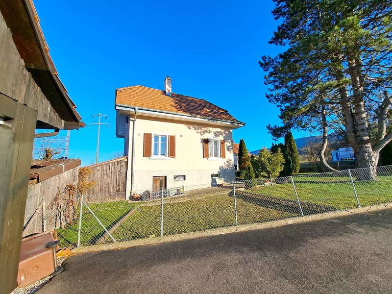 2-story house, brown roof, beige exterior, windows with brown shutters, fenced yard, chain-link fence, grass, trees, clear sky, mountains in the distance