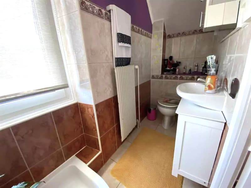 Bathroom with a sink, toilet, radiator, towels, window, tiles, and a trash can.
