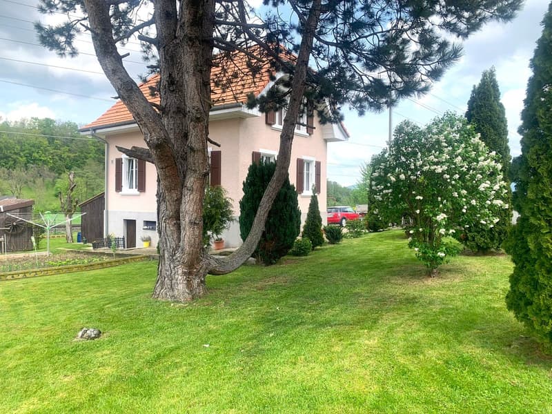 2 story house, beige color, orange roof, multiple windows, small garden, grassy area, shrubbery, trees, car parked outside