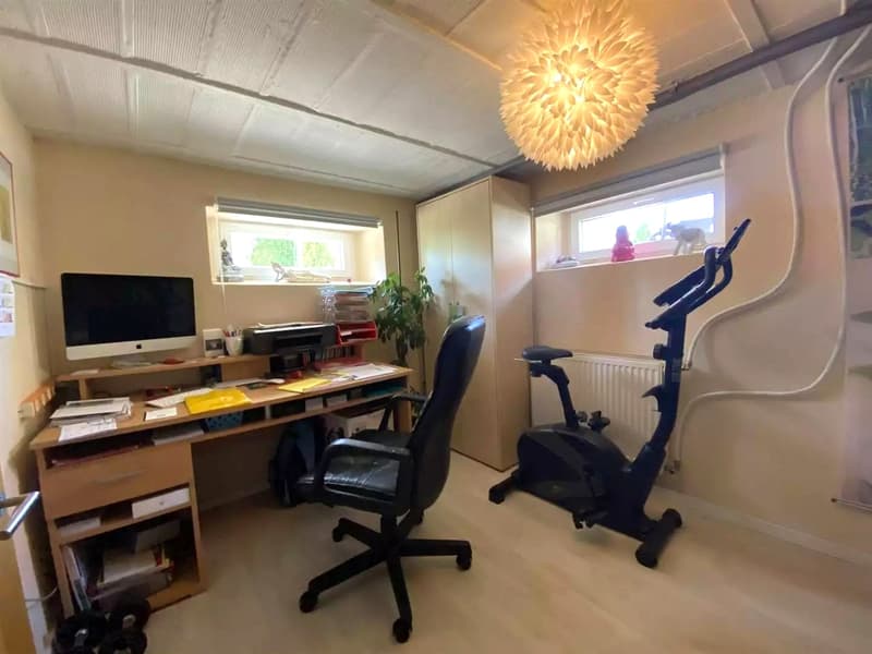 A room with a wooden desk, a computer monitor, a printer, a chair, and a potted plant. The room has a lamp and a large window with a view outside. There is also a treadmill, a cabinet, and a radiator in the room.