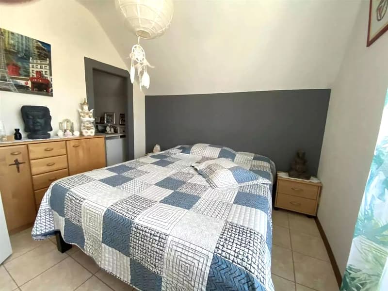 a double bed with a quilted cover, a nightstand with drawers, wooden cabinets, a pendant light, a wall with a gray accent, and various decorative items
