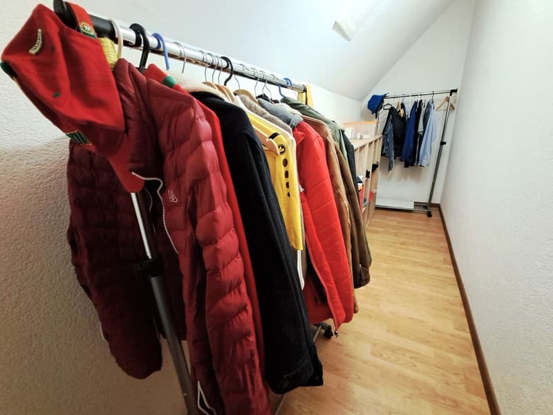 Room with hanging clothes and wooden floor. Storage box and additional hangers visible. Wall with light. Warm color lights up the room.
